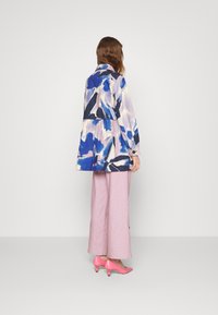 Nainen seisoo selkä kameralle, päällään pitkähihaisessa abstraktissa sinivihreässä ja vaaleanpunaisessa kuosissa olevassa takissa, vaaleanpunaisissa leveälahkeisissa housuissa ja vaaleanpunaisissa kengissä.