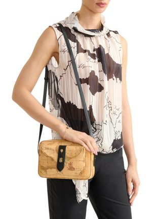 Donna che indossa una camicetta senza maniche con stampa di mappe e pantaloni neri, che tiene una borsa a tracolla color cuoio con un design di mappa vintage e tracolla nera.