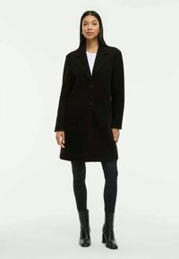 Schwarzer maßgeschneiderter Mantel mit Reverskragen, einzelnem Knopfverschluss und Seitentaschen; kombiniert mit schwarzen Leggings und Stiefeletten.