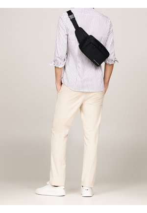 Homme portant une chemise rayée, un pantalon beige, des baskets blanches et un sac bandoulière noir dans le dos sur un fond uni.