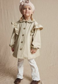 Beige kinderregenjas met gerimpelde kraag en mouwen, met grote knopen. Draag het over witte broek met bloemenpatroon en bruine schoenen.