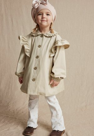 Beige kinderregenjas met gerimpelde kraag en mouwen, met grote knopen. Draag het over witte broek met bloemenpatroon en bruine schoenen.