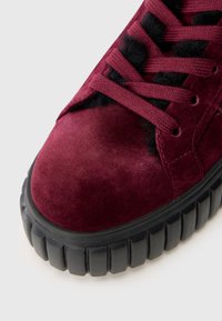Burgunderfarvet ruskinds sneaker med teksturerede sorte accenter, rund tå og tyk, skridsikker sort gummisål. Snørebåndene er i matchende burgunder.