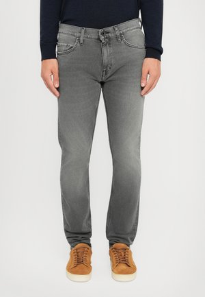 Mand iført slim-fit grå jeans og brune ruskindssneakers, stående foran en ensfarvet hvid baggrund.