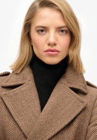 Brązowy płaszcz dwurzędowy w jodełkę z szerokimi klapami, noszony na czarnym prążkowanym golfie, o gładkiej fakturze i strukturalnej sylwetce.