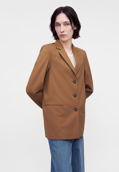 Personne aux cheveux courts et foncés portant un blazer marron boutonné sur un haut clair et un jean bleu, debout les mains derrière le dos.