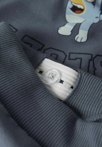 Poignet de manche côtelée bleue avec un élastique blanc réglable à l'intérieur, faisant partie d'un vêtement arborant un design de personnage animal de bande dessinée et un texte en bloc.