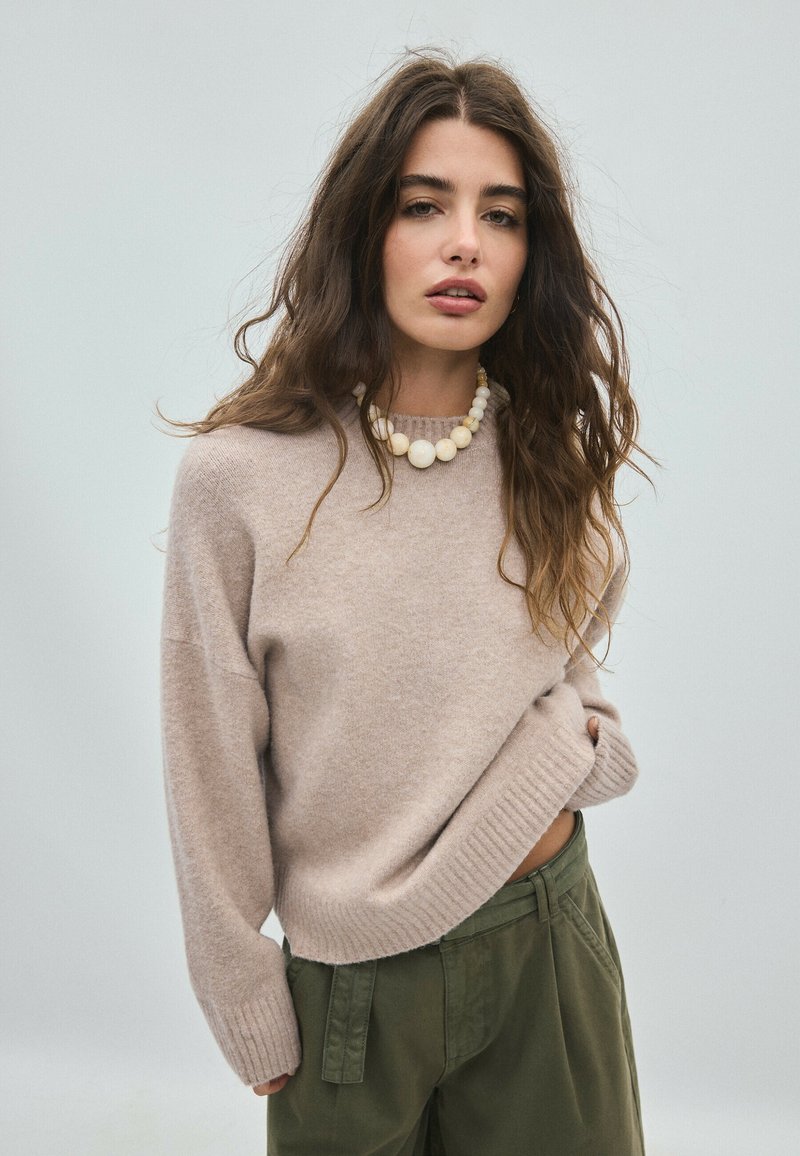 Jeune femme aux longs cheveux ondulés portant un pull beige, un grand collier de perles et un pantalon vert, posant devant un fond uni gris clair.