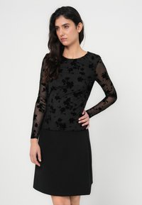 Μαύρο μακρυμάνικο τοπ με λουλουδάτο μοτίβο σε διαφανές ύφασμα, συνδυασμένο με απλή μαύρη φούστα. Απλός λαιμός και λεία υφή.