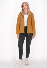 Mosterdgele waterproof jas met een capuchon, ritssluiting aan de voorkant en zijzakken, gecombineerd met zwarte skinny jeans en lichtgekleurde sneakers.