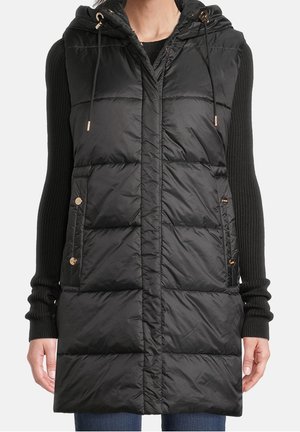 Woman wearing sleeveless black padded hooded vest over a black long-sleeve ribbed sweater with blue jeans.