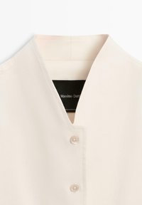 Gros plan d'un chemisier couleur crème à col en V avec deux boutons et une étiquette noire visible à l'intérieur du col portant l'inscription « Massimo Dutti ».