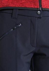 Pantalon bleu marine avec poche zippée sur le côté et fermeture par bouton, accompagné d'une veste rouge texturée avec fermeture éclair frontale.