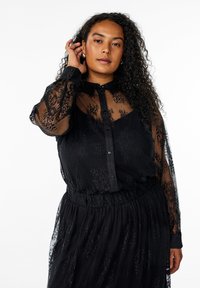 Black lace blouse with sheer long sleeves, front button closure, and a textured floral pattern. Loose fit and gathered waist detail.