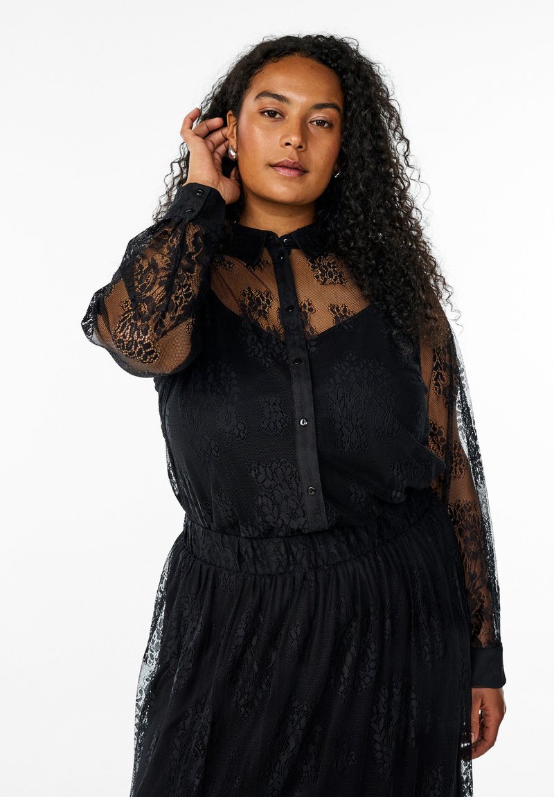 Blouse en dentelle noire avec des manches longues transparentes, fermeture à boutons à l'avant et un motif floral texturé. Coupe ample et détail de taille froncé.