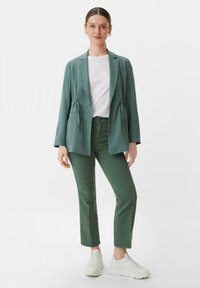 Zielony żakiet z regulowanymi sznurkami w pasie, zestawiony z białą koszulką i zielonymi spodniami. Modelka nosi białe trampki. Gładki materiał, luźny krój.