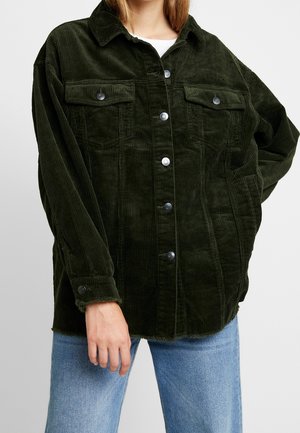 Personne portant une veste en velours côtelé vert foncé boutonnée avec des poches poitrine à rabat et un jean bleu clair sur un fond uni.