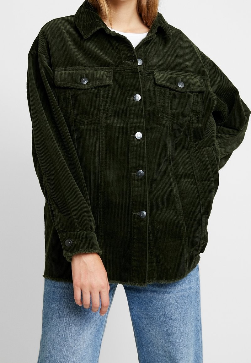 Personne portant une veste en velours côtelé vert foncé boutonnée avec des poches poitrine à rabat et un jean bleu clair sur un fond uni.