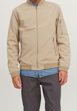 Giacca bomber beige in nylon. Presenta una zip frontale, tasche laterali e polsini a coste. Indossata sopra una camicia di denim e pantaloni grigi.