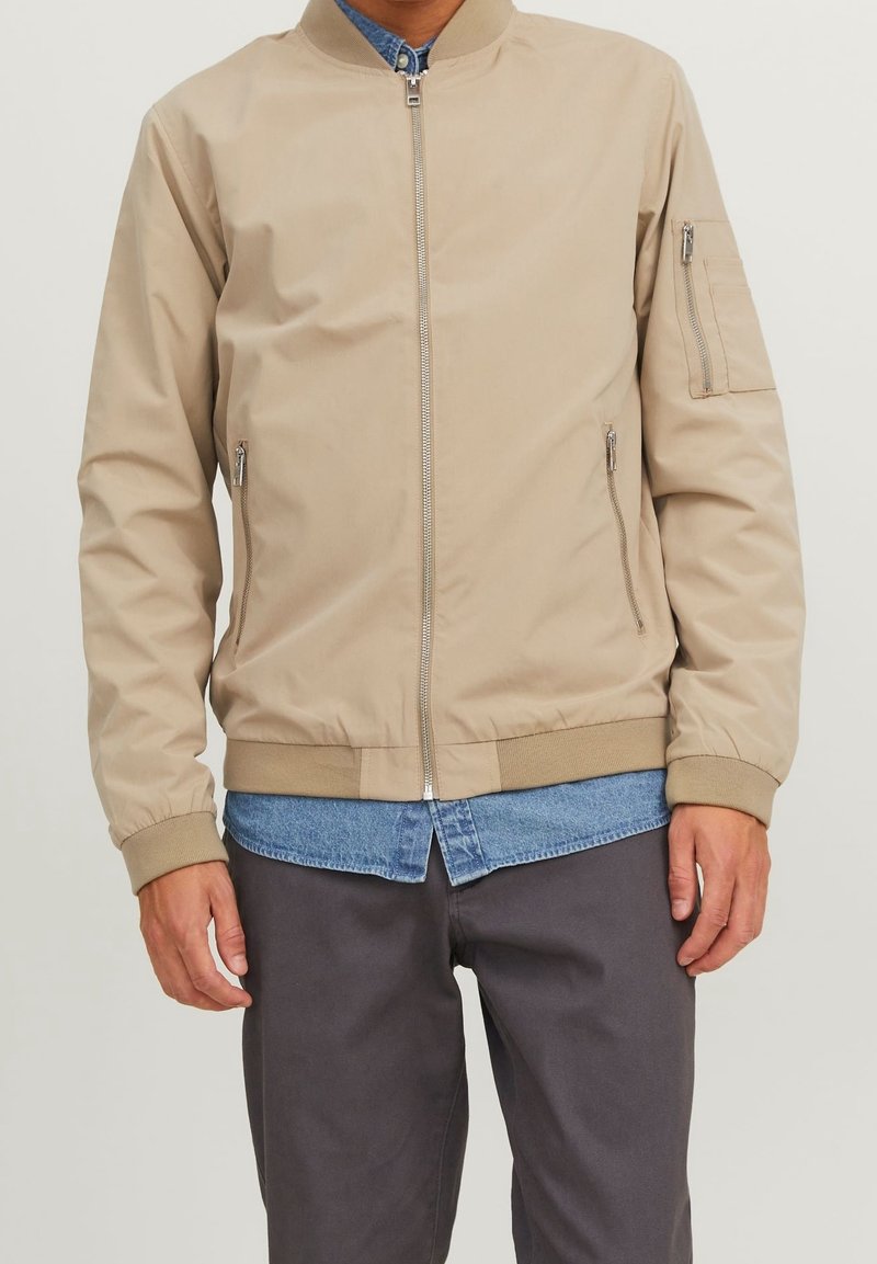 Beige bomberjacka i nylon. Har en framzipper, sidofickor och ribbade ärmslut. Bärs över en denimskjorta och gråbyxor.