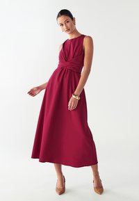 Burgundska haljina bez rukava s uvijenim korzetom, lepršavom suknjom i glatkom teksturom. Nosiva sa zlatnom narukvicom i uzorkovanim potpeticama.