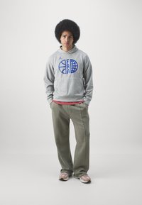 Felpa grigia con logo blu, camicia rosa sotto, pantaloni verde oliva e scarpe beige con accenti chiari. Il modello si trova su uno sfondo neutro.