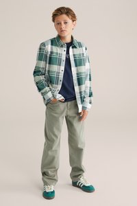 Raiba krekls zaļā un baltā krāsā, virsū tumši zila T-krekla, apvienots ar gaiši zaļām biksēm un bērza zaļām kedām ar baltām svītrām. Standarta piegriezums.