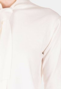 Top color crema con tessuto morbido e testurizzato. Presenta un dettaglio a fiocco annodato al collo e una silhouette aderente.