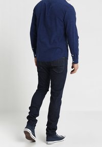 Marinblå rutig långärmad skjorta, mörkblå slim-fit jeans och marinblå sneakers med vita detaljer, sedd från baksidan.