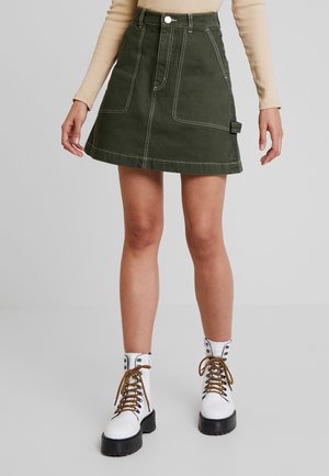 Vrouw draagt een groene A-lijn rok met voorzakken en witte veterlaarzen met zwarte zolen, staand tegen een effen achtergrond.