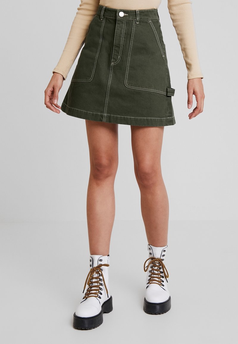 Femme portant une jupe trapèze verte avec des poches frontales et des bottes blanches à lacets avec semelles noires, debout devant un fond uni.