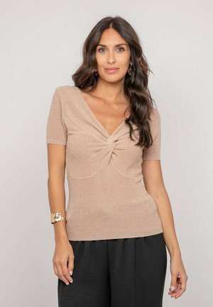 Femme aux longs cheveux ondulés et foncés porte un haut beige en maille à manches courtes avec un détail torsadé à l'avant et un pantalon noir, posant devant un fond uni.