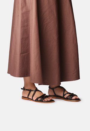 Persoon draagt een bruine enkellange rok en zwarte platte sandalen met bandjes en gesp op een witte achtergrond.