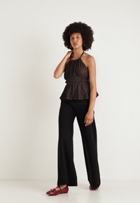 Haut à col halter marron foncé avec taille peplum, en tissu texturé ; associé à un pantalon noir évasé et des chaussures rouges, se tenant devant un fond clair.