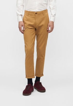 Bărbat purtând pantaloni chino de culoarea muștarului rulați, șosete negre, adidași burgundy și o cămașă albă cu mâneci lungi, stând în fața unui fundal simplu.