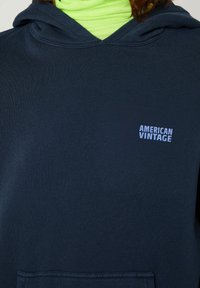 Jūras spēku kapuce no strukturēta auduma, ar priekšējo kabatu un mazu balto "American Vintage" logotipu uz krūtīm.