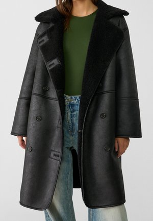 Vrouw draagt een oversized zwart schapenvachtjas met brede revers, donkergroen topje en lichtblauwe jeans, staand tegen een effen achtergrond.