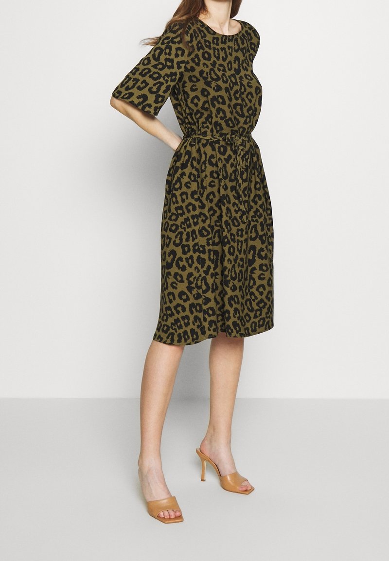 Femme portant une robe mi-longue kaki avec imprimé léopard noir, manches mi-longues et ceinture nouée à la taille, associée à des sandales à talons hauts couleur beige.
