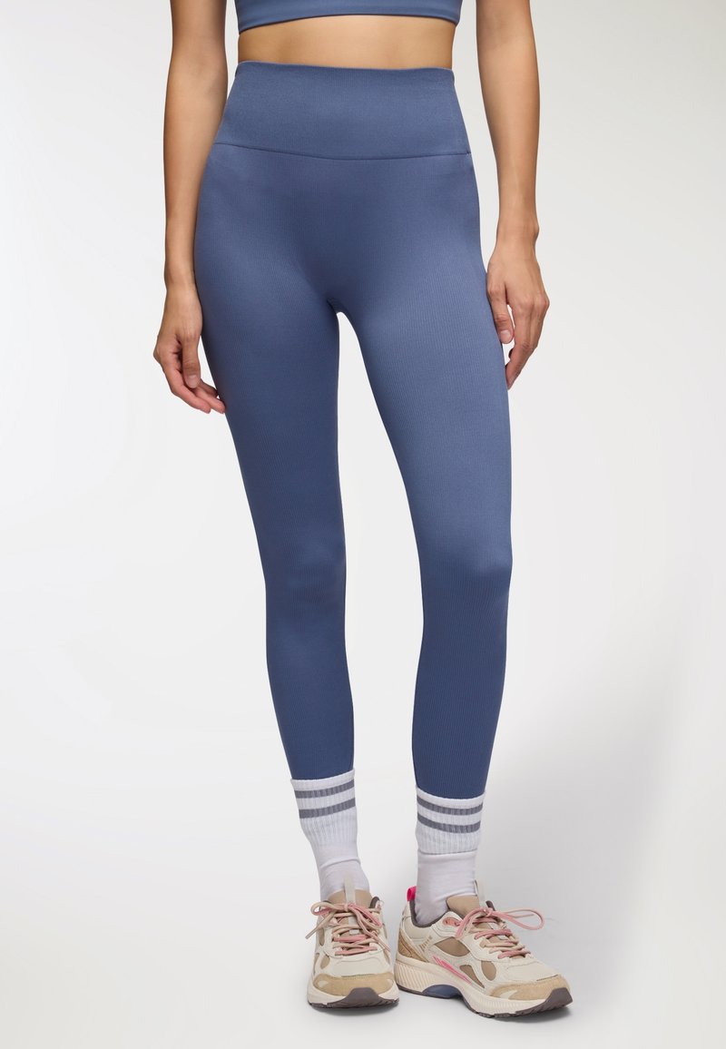 Legginsy w kolorze niebieskim, z wysokim stanem i prążkowaną fakturą, wykonane z elastycznego materiału. W połączeniu z jasnymi sneakersami i białymi skarpetkami w szare paski.