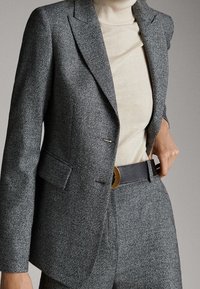 Blazer gris texturé avec revers crantés, fermeture à un bouton et poches à rabat, associé à un col roulé crème et une ceinture noire.