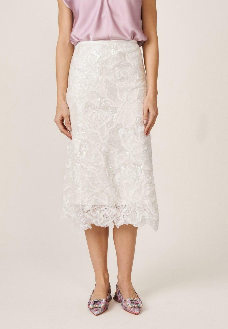 Woman wearing a white lace midi skirt with floral patterns, a light pink sleeveless top, and patterned pointed-toe heels.