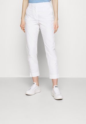 Persoon die witte korte broeken en witte platformsneakers draagt, staand tegen een effen lichte achtergrond.