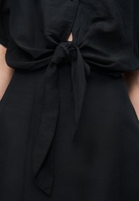 Μαύρο ύφασμα δεμένο με κόμπο μπροστά πάνω σε μια ταυτόχρωμη ψηλόμεση βάση, που αφήνει μια μικρή οπτική στο μέσο του κορμού κάτω από τον κόμπο.