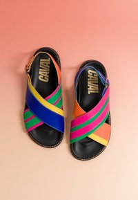 Paire de sandales noires avec des brides croisées multicolores et des boucles à la cheville, nom de la marque "CANAL" en or sur la semelle, sur fond pêche.