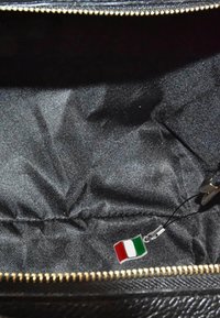 Zwart interieur van een tas met een gouden rits, voorzien van een klein Italiaans vlaggetje als hanger aan een zwarte riem. Gladde, textiele structuur.