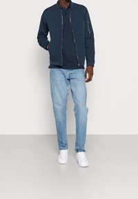 Muž v námořnické bomber bundě přes námořnickou košili s puntíky, světlých modrých džínách a bílých teniskách stojí na jednobarevném pozadí.