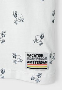 Hvid t-shirt med sorte illustrationer af skateboarding hunde, med et vævet mærke der står "VACATION SCRAPBOOK AMSTERDAM" i kraftige farver.