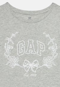 Ανοιχτό γκρι ύφασμα με λευκό στεφάνι από λουλούδια, σχέδιο φιόγκου και το κείμενο "GAP" πάνω από το "Est. 1969" τυπωμένο με λευκά γράμματα.
