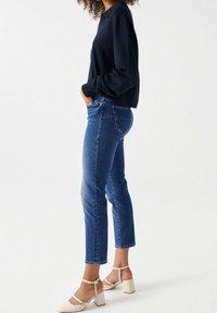 Svart långärmad topp, figurnära blå jeans och vita sandaler med blockklack. Slät tyg; klassisk denimdesign; rak passform med sidofickor.