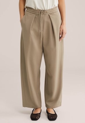 Nő bézs színű, széles szárú nadrágban, amelynek elasztikus, zsinóros derékrésze van, fekete horgolt lapos cipőkkel, sima háttér előtt áll.